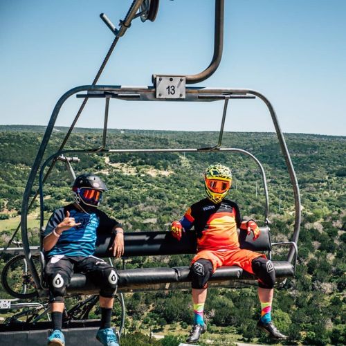 Two mountain bikers on a chairlift, wearing helmets and colorful gear, with a scenic green landscape in the background.