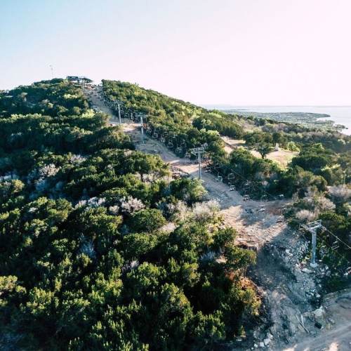 Aerial view of a hillside with a ski lift, surrounded by lush greenery and a glimpse of water in the distance.