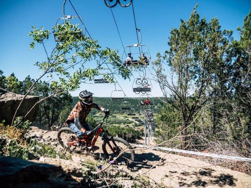 A mountain biker descends a trail with ski lifts in the background, surrounded by trees and a clear blue sky.