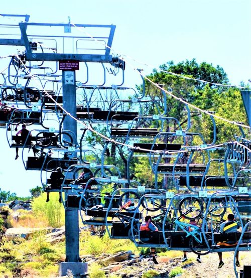 A ski lift with empty chairs and a few riders, surrounded by trees and a clear blue sky.