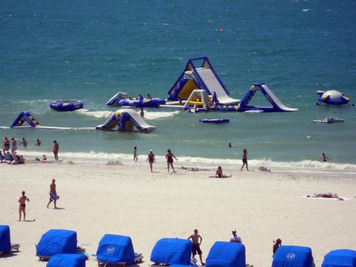 A beach scene with inflatable water attractions, people swimming, and blue cabanas on the sand.
