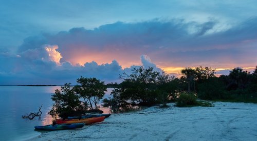 A serene beach at sunset with colorful clouds, kayaks on the shore, and lush greenery along the water's edge.