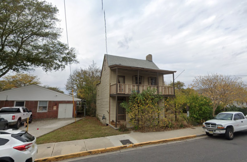 A two-story house with a porch, surrounded by trees, and a garage nearby on a quiet street.