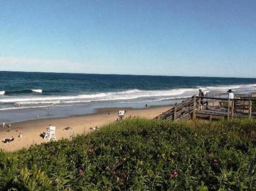 A sunny beach scene with waves, people enjoying the sand, and a wooden walkway surrounded by greenery.