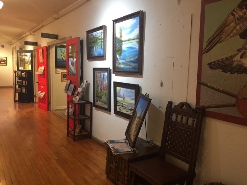 A gallery hallway featuring framed paintings, a wooden chair, and a red door, with wooden flooring and display stands.