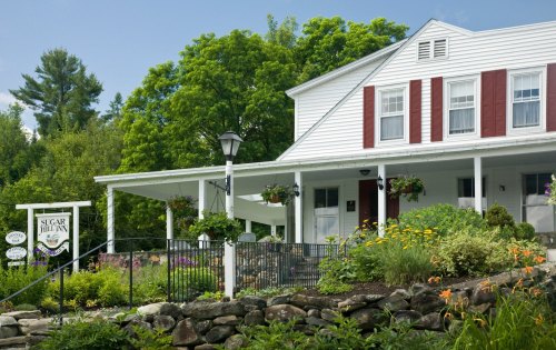 A charming white house with red shutters, surrounded by lush greenery and colorful flowers, featuring a welcoming sign.