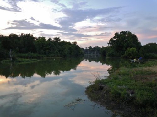 A serene river scene at sunset, reflecting trees and clouds, with a small boat near the shore.