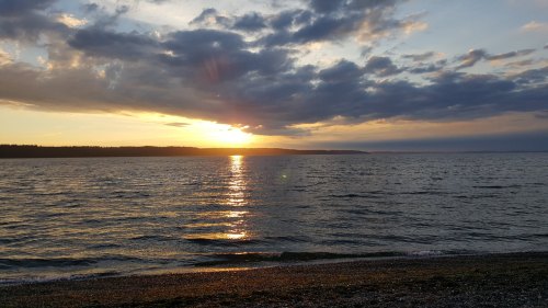 Sunset over a calm sea, with clouds reflecting golden light and a shoreline in the foreground.
