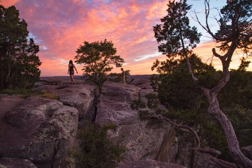 A person walks on rocky terrain at sunset, with vibrant clouds and trees in the background.
