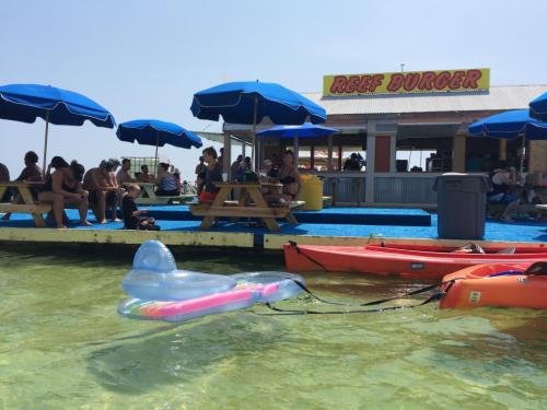 A beach scene with a burger stand, people sitting under blue umbrellas, and colorful kayaks on clear water.