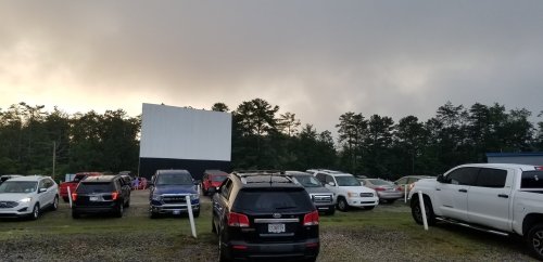 A drive-in theater with cars parked in front of a large screen, surrounded by trees and a cloudy sky.