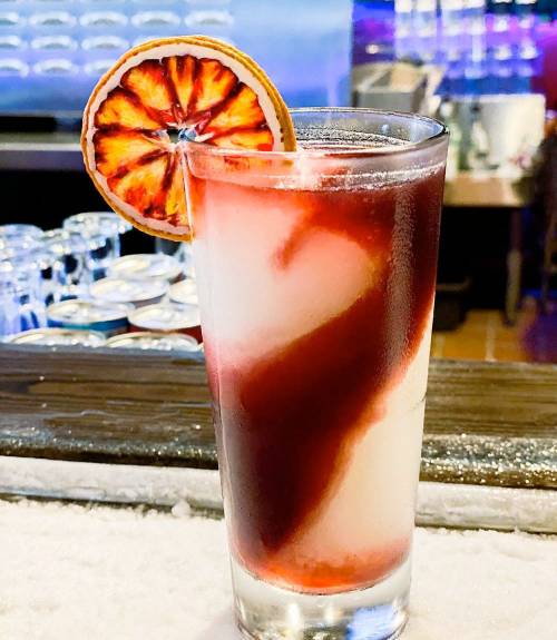 A colorful cocktail with a swirl of red and white, garnished with a dried orange slice, served in a tall glass.