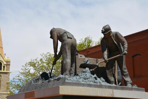 Statue of two miners working, with tools, set against a blue sky and historic buildings in the background.