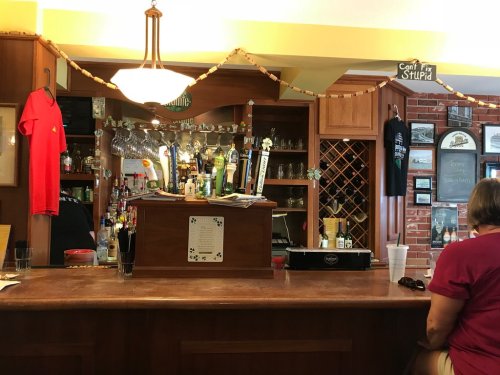 A cozy bar interior with a wooden counter, beer taps, and shelves of drinks, featuring a patron sitting at the bar.