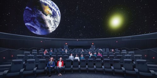 A planetarium with an audience seated, featuring a large Earth and sun projected on the dome above.