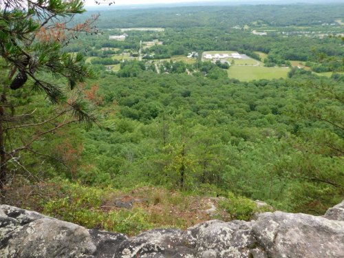 A scenic view from a rocky overlook, showcasing a lush green valley and distant fields under a cloudy sky.
