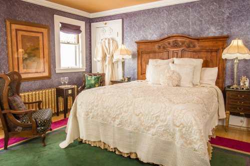 Cozy bedroom with a large bed, elegant decor, and vintage furniture against a purple wallpaper backdrop.