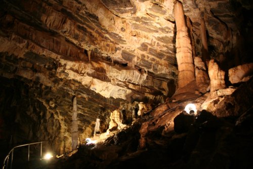 Stalactites and stalagmites in a dimly lit cave, with stone formations and artificial lights illuminating the scene.