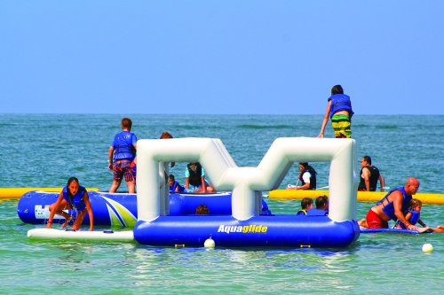 Children and adults play on inflatable water structures in a sunny beach setting.