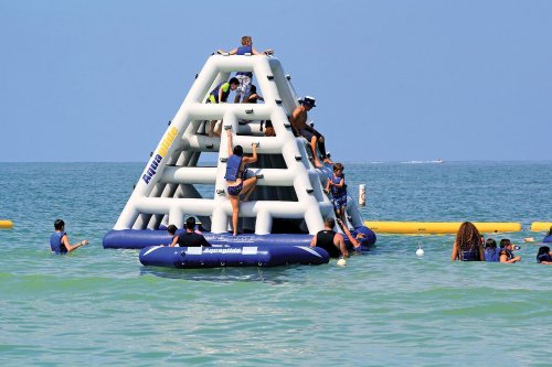Children and adults climb an inflatable water structure in a sunny ocean, with others playing nearby.