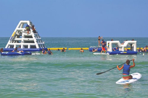 A lively beach scene with people enjoying inflatable water activities and paddleboarding in clear blue water.