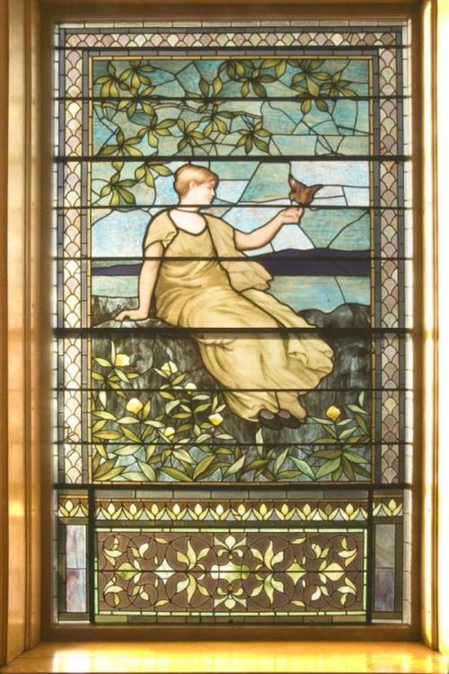 Stained glass window featuring a woman in a yellow dress holding a bird, surrounded by greenery and floral patterns.