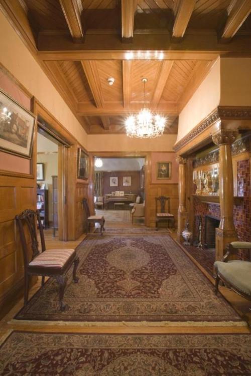 A spacious hallway with wooden beams, ornate decor, and a chandelier, featuring chairs and a patterned rug.