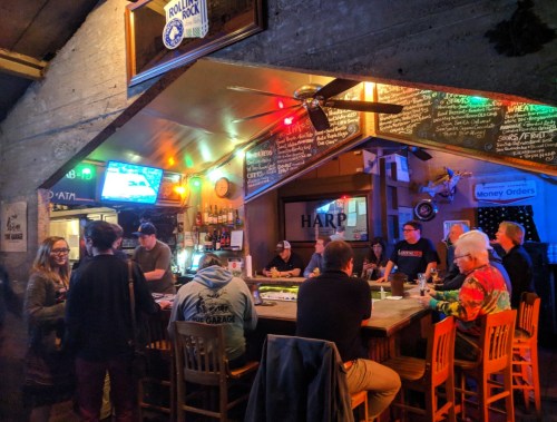 A cozy bar scene with patrons at the counter, colorful decor, and a TV showing a game in a warm, inviting atmosphere.