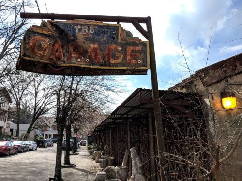 Weathered sign reading "Garage" hangs above a rustic building, with trees and parked cars lining the street.