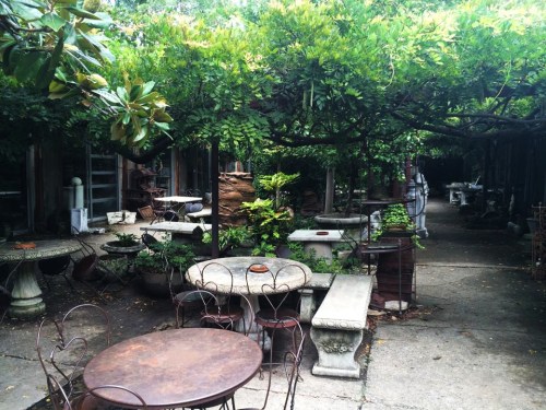 A serene garden space with stone tables, chairs, and lush greenery overhead, creating a tranquil atmosphere.