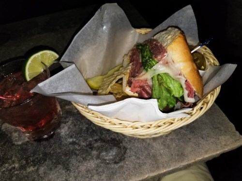 A sandwich with lettuce and cheese in a basket, accompanied by pickles and a drink with a lime wedge.