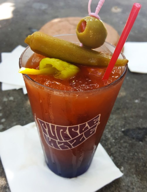 A tall glass of a red cocktail garnished with an olive, pickled green bean, and pepper, served on a napkin.