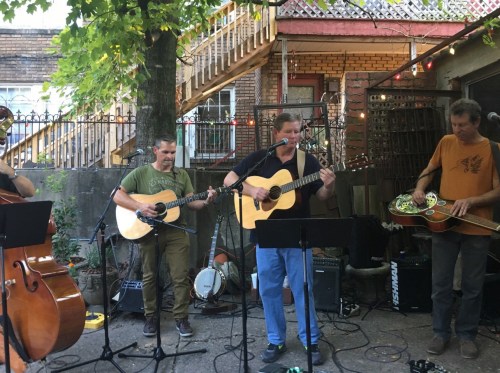 A group of musicians playing acoustic instruments outdoors, surrounded by greenery and a rustic setting.