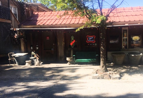 A rustic building with a red-tiled roof, green bench, and neon signs, surrounded by trees and outdoor seating.