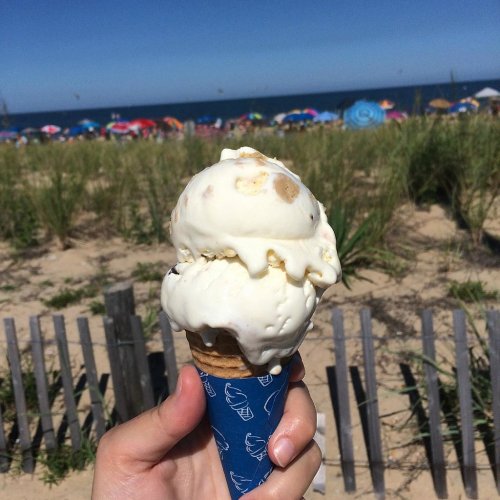 A hand holds a double scoop ice cream cone against a beach backdrop with umbrellas and tall grass.