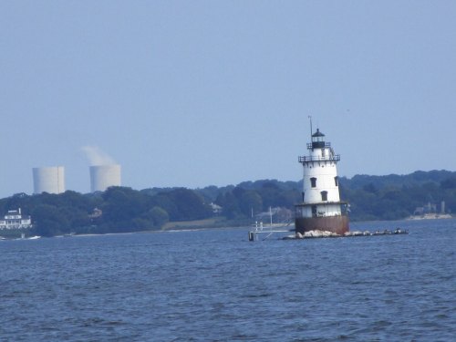 A lighthouse stands on a rocky outcrop in the water, with power plant cooling towers visible in the background.