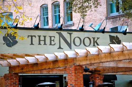 Sign for "The Nook" restaurant with a wooden pergola in front and brick walls, set in a charming urban environment.