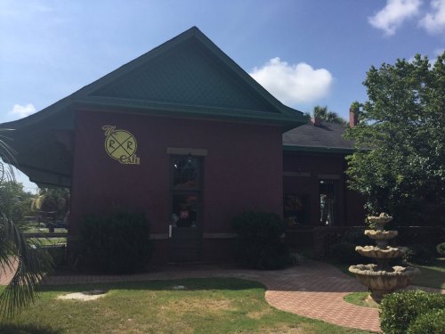 A charming café building with a green roof, surrounded by greenery and a fountain in front, under a clear blue sky.