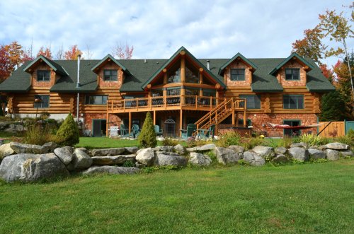 A large log cabin with multiple gables, surrounded by greenery and rocks, under a partly cloudy sky.