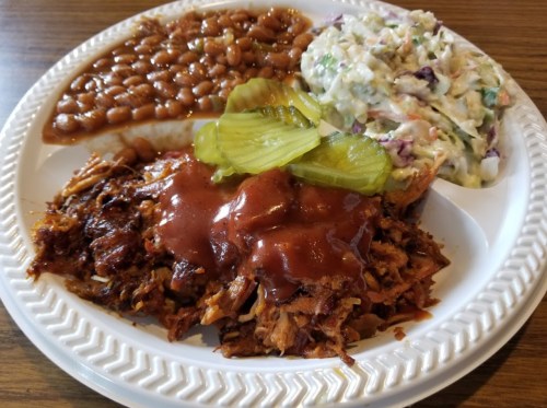 A plate of pulled pork topped with barbecue sauce, pickles, baked beans, and coleslaw.