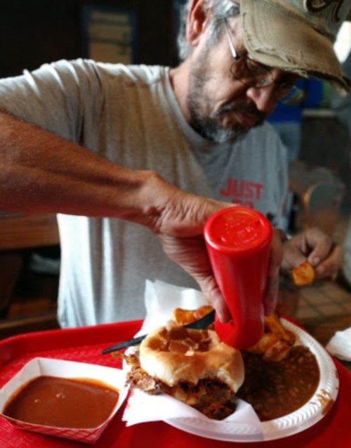 A man in a cap squeezes ketchup onto a sandwich and fries on a red tray, with a side of sauce.
