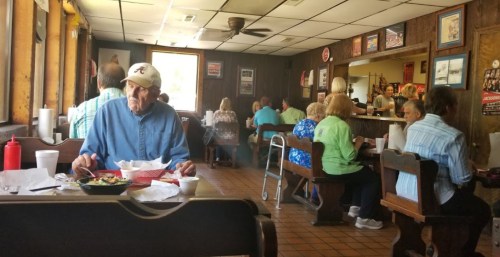 A busy diner with patrons eating at tables, an elderly man in a cap enjoying a meal in the foreground.