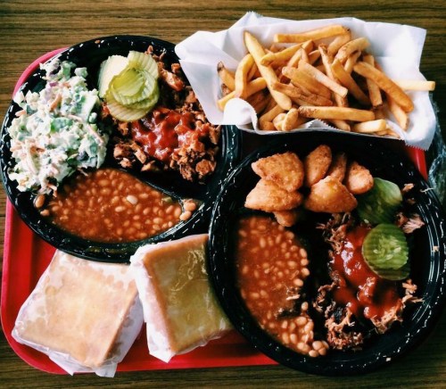 A red tray with two plates of pulled pork, baked beans, coleslaw, pickles, fries, and two pieces of cornbread.