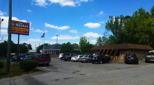 A parking lot with several cars outside a restaurant called "The Rocket," under a blue sky with fluffy clouds.