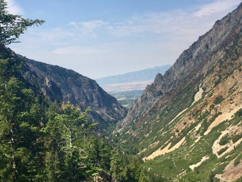A scenic view of a valley between steep mountains, with lush greenery and a distant landscape under a blue sky.
