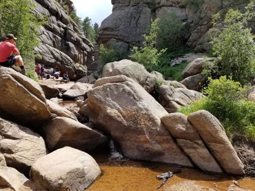 A rocky landscape with large boulders and a small stream, surrounded by greenery and people sitting in the background.