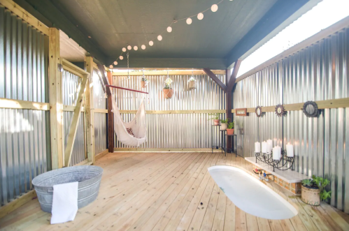 Cozy outdoor space with wooden flooring, a hammock, potted plants, and string lights, featuring a bathtub and candles.
