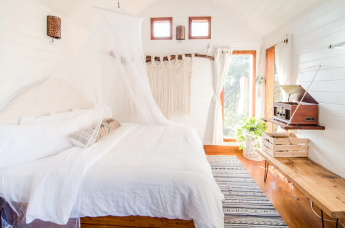 Cozy bedroom with a white bed, wooden accents, and natural light, featuring a vintage record player and plants.