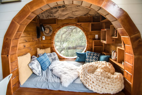 Cozy wooden nook with a circular window, plush pillows, and a chunky knit blanket, surrounded by nature.