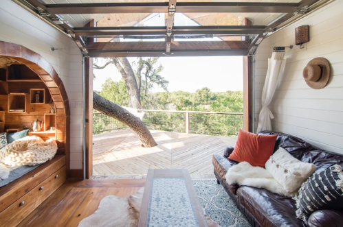 Cozy interior of a tiny home with a large window, wooden accents, and a view of a deck surrounded by trees.